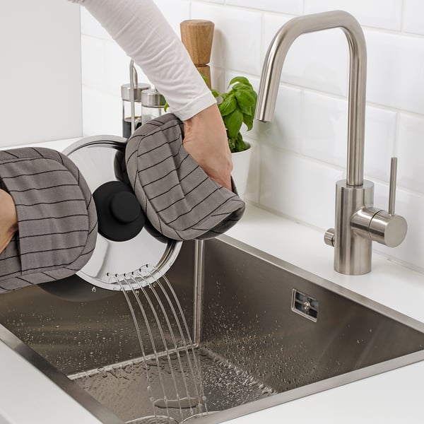 Person washes herbs in sink using strainer, with modern tap, white tiles in background.