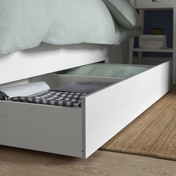Bed with open storage drawer under HEMNES frame, showing folded blankets and sheets.