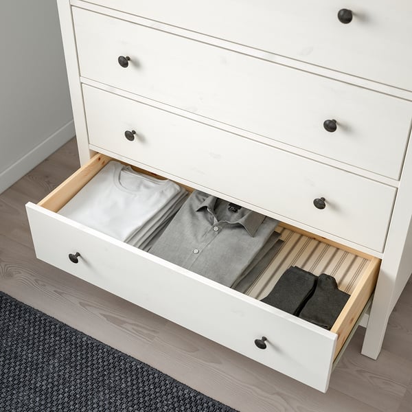 White wooden dresser with four drawers, three closed, one open revealing folded clothes and socks, anchored for safety.