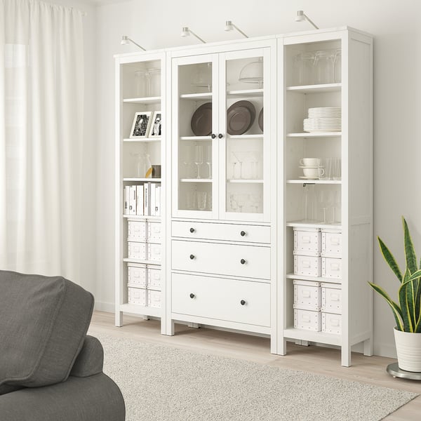 White HEMNES storage unit with glass doors, shelves, and drawers next to a grey sofa, displaying glassware, dishes, and books.