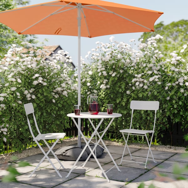 Small outdoor setup with orange umbrella, white table, and two chairs surrounded by blooming bushes.