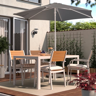 An outdoor patio with a grey HÖGÖN parasol, white metal and light wood dining set, surrounded by potted plants.