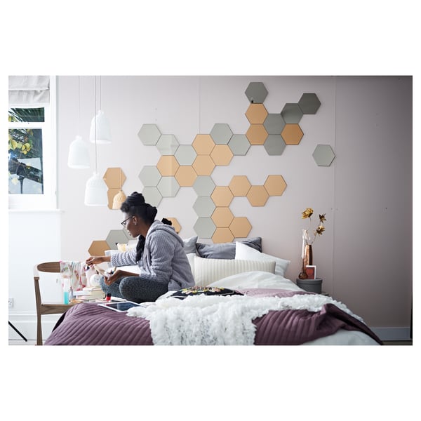 A person sits on a bed with white blankets against a wall adorned with hexagonal mirrors in grey and tan, creating a modern, calm bedroom scene.