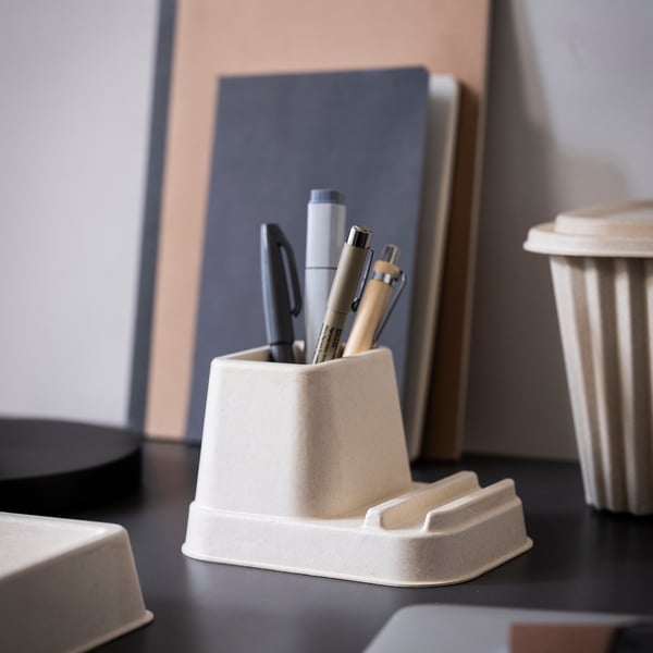 A small, beige HÖNSNÄT organiser holding pens on a desk. Its compact, made from moulded paper pulp, and fits well among other desk items.