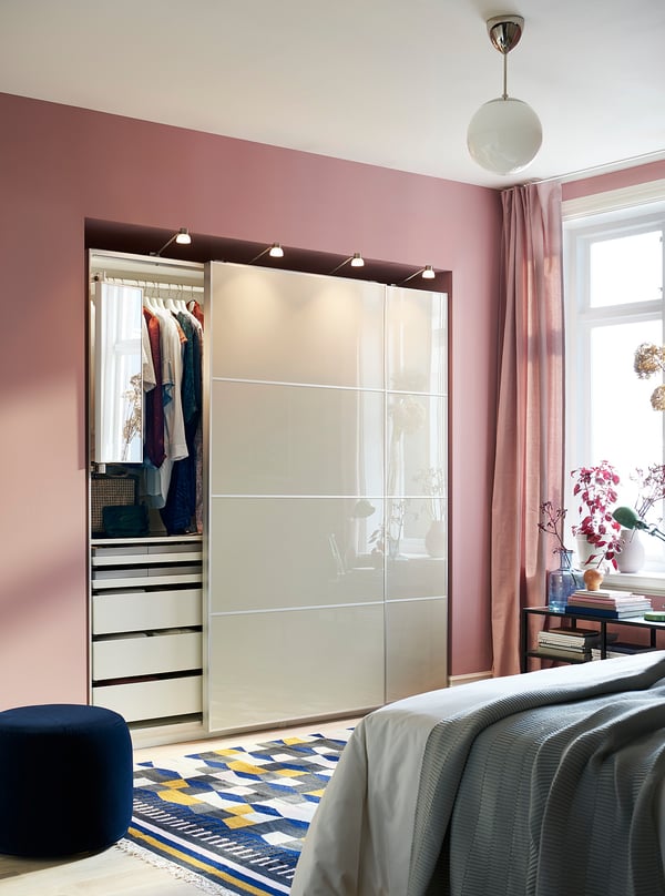 Bedroom with HOKKSUND pinkish wardrobe, mirrored doors. Bed with grey blankets, divine rug, round blue stool.