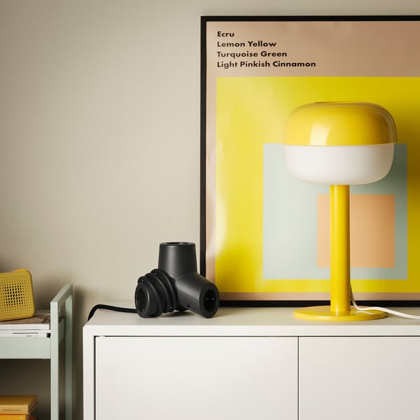 Black HORNFELS 4-way extension on white shelf. Near yellow lamp, colorful art, wall plug.