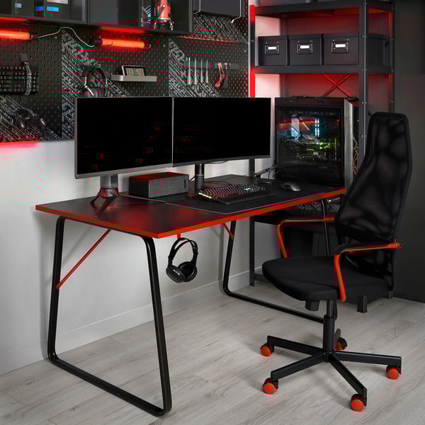 HUVUDSPELARE gaming setup with dual monitors, keyboard, and tower on red-black desk. Mesh chair fits underneath. Tech accessories on shelves.