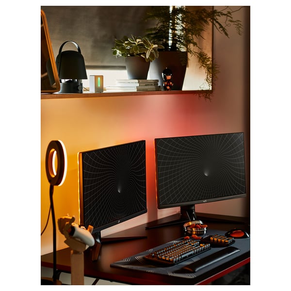 A black gaming desk setup with dual monitors, ergonomic keyboard, ring light, and plant decorations.
