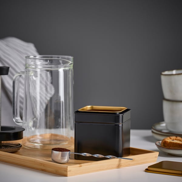 A wooden tray with a metal coffee scoop, a black container, a glass pitcher, and mugs. The scoop has a long handle and is filled with coffee grounds.