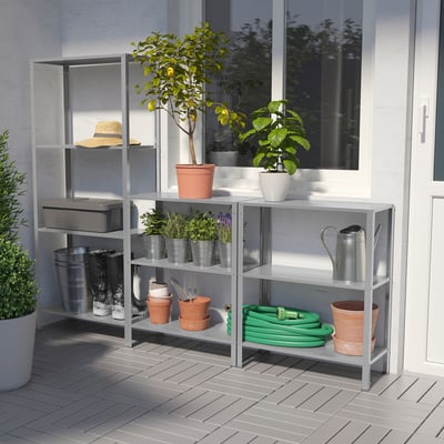 Silver shelving unit with plants, tools outdoors against white wall.