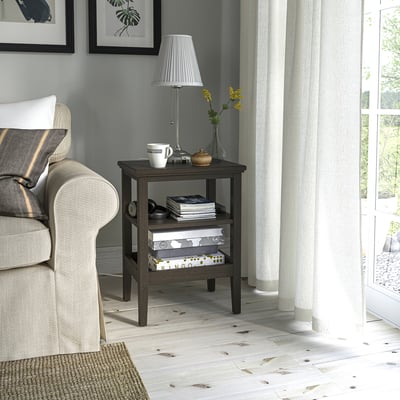 IDANÄS side table next to grey chair, holding lamp, mugs, books. Dark wood, white floors. Ideal for tight spaces.