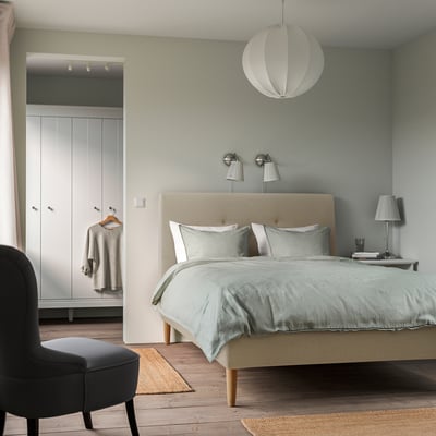 Bedroom with IDANÄS bed, blue linens, pendant lamp, black chair on rug, white wardrobe. Calm, minimalist design.