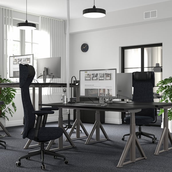 Modern office with black IDÅSEN desks, high chairs, multi-monitor setups, hanging lamps, and plants.
