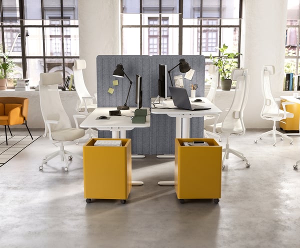Modern office with IDÅSEN yellow storage units, white desks, chairs, and grey partitions. Large windows, plants, and orange accent chairs.