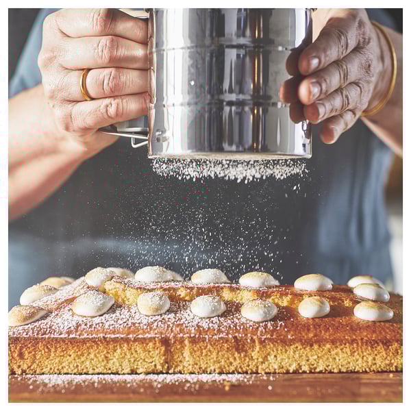 Two hands use an IDEALISK sifter, silver in colour, to dust white icing sugar onto a large rectangular baked good topped with meringues.