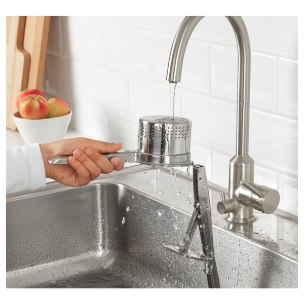 Silver tap pouring water into stainless steel sink with hand holding strainer, beside bowl of apples.