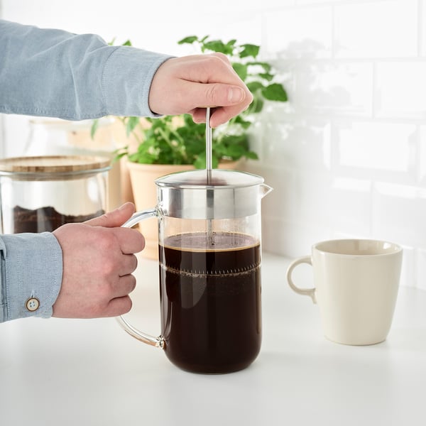 A person is pressing a plunger in a glass coffee maker, extracting coffee into a jug. A white mug is beside the maker.