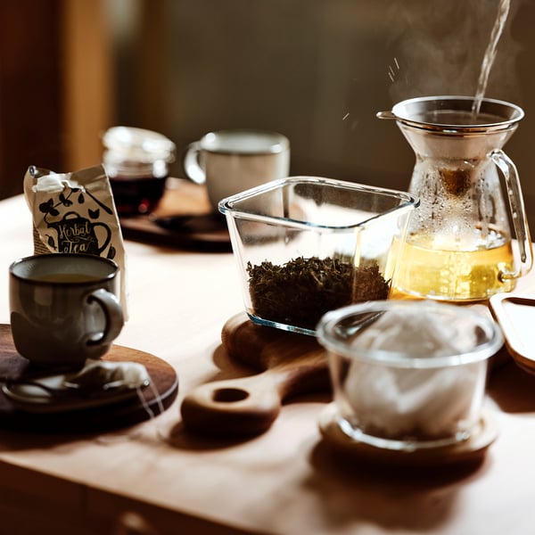 Glass food container with loose tea and infuser, placed on table with mugs.