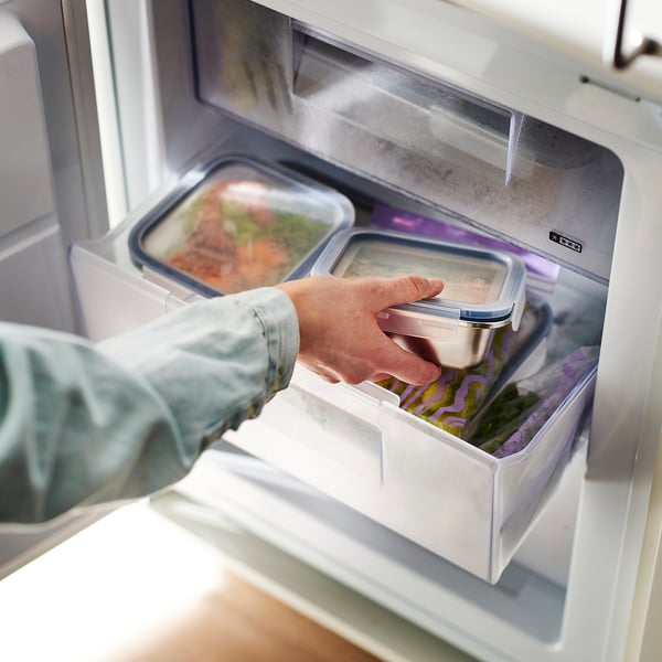 Person placing a stainless steel food container with a lid into a fridge. The container is transparent and designed for storing leftovers.