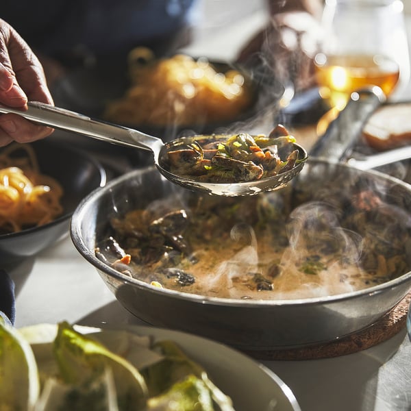 A hand holds tongs placing sautéed vegetables from a frying pan into a bowl, steam rises, showing even heat distribution.