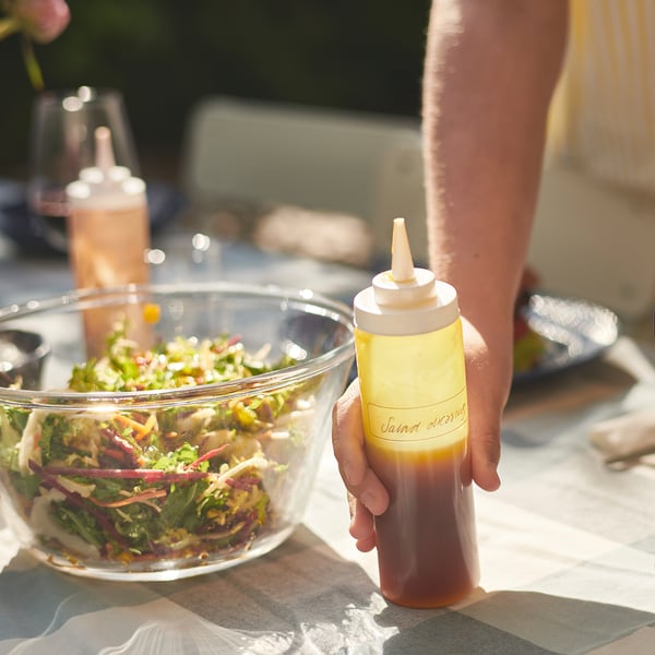 Hand holding yellow IKEA 365+ squeeze bottle with label salad dressing over bowl of salad.