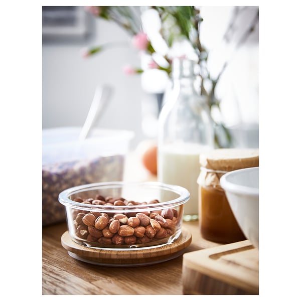 Clear round glass container filled with hazelnuts, on a wooden table, surrounded by jars and flowers.