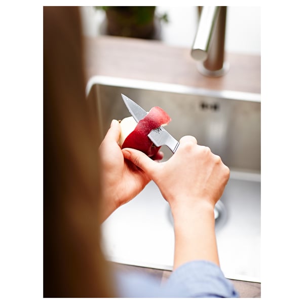 Hands peeling red apple with small knife near sink.