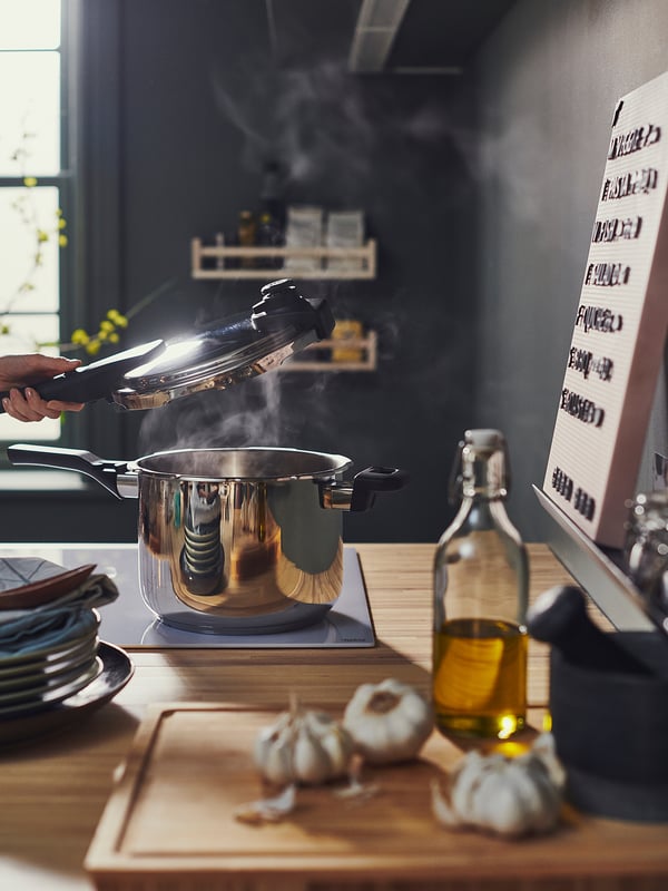 A modern kitchen scene with IKEA 365+ stainless steel pressure cooker on a wooden countertop with garlic, olive oil, and stacked plates.