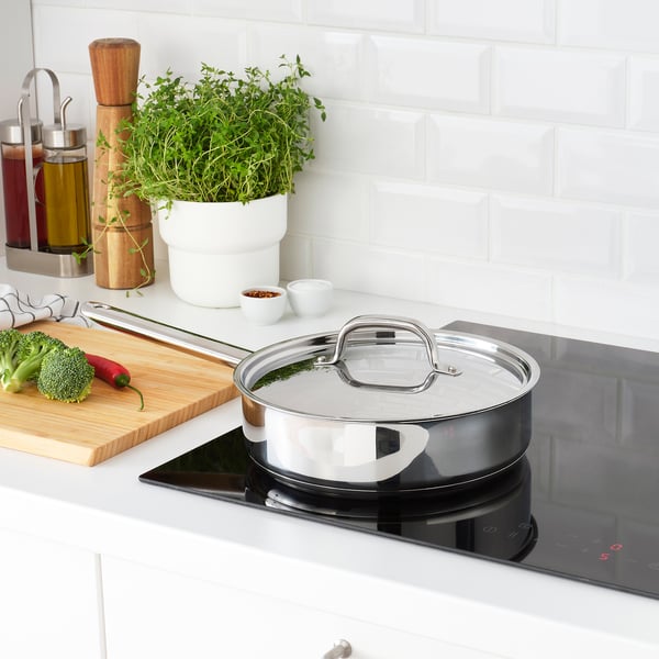 Modern kitchen with stainless steel pan on induction stove, cutting board with veggies, condiments, and a plant.