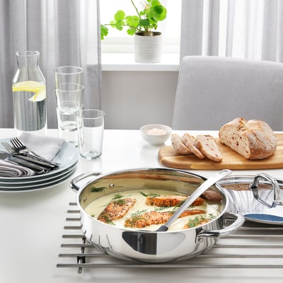 Stainless steel sauté pan on trivet with grilled fish, bread, and glasses on dining table by window.