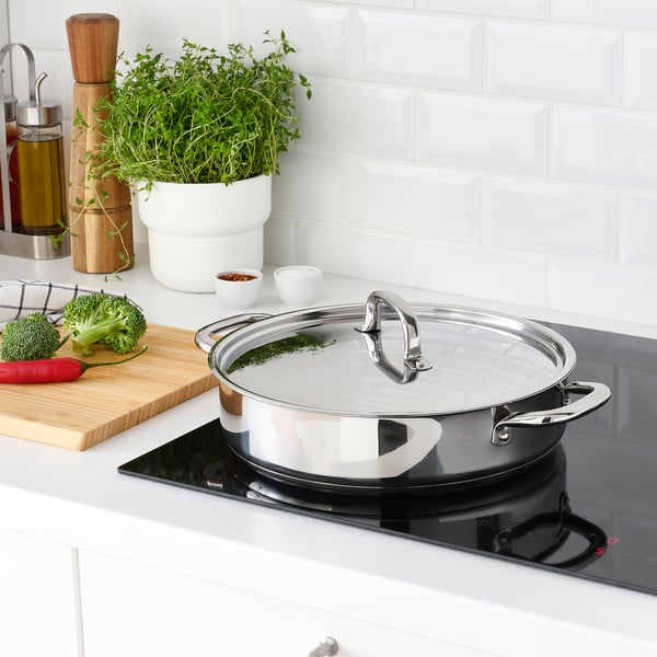Modern kitchen with induction stove, featuring a covered stainless-steel sauté pan, fresh herbs, and chopped vegetables on a cutting board.