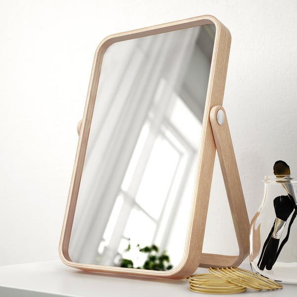 Mirror on table with wooden stand, reflecting room. Beside makeup brushes and coiled cords.