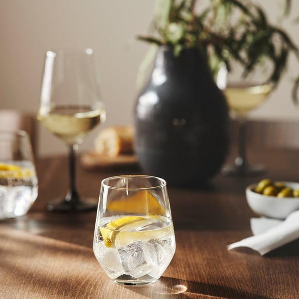 On a wooden table sits a clear wine glass with a lemon slice, next to a vase of flowers and a bowl of olives.