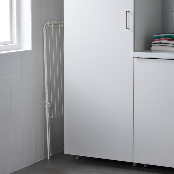 A white drying rack, slim, folds beside cabinet. Tall, indoor/outdoor, compact when not used.