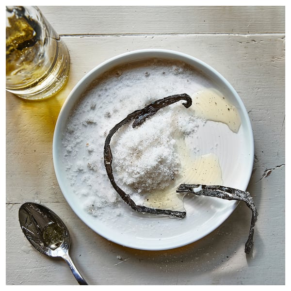 A white plate with vanilla beans and sugar, a spoon, and a glass container of oil beside it.