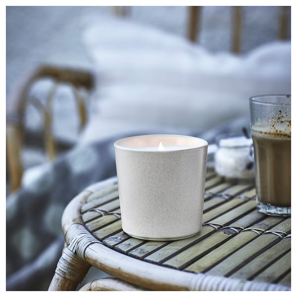 A lit white JÄMLIK scented candle sits on a wicker table outside, beside a cup of coffee.