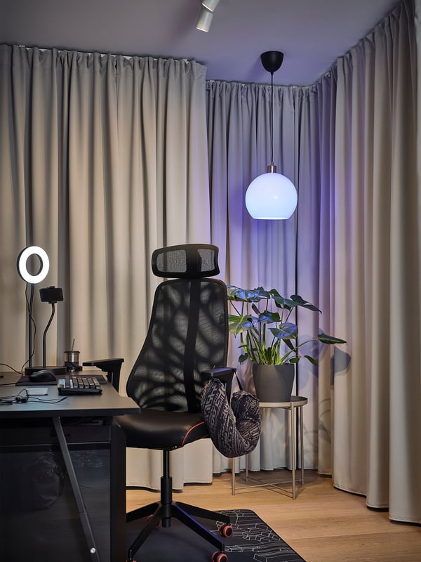 Black home office with desk, lamp, chair, ring light, plant, grey curtains.