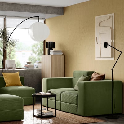 Modern living room: green corduroy sofa, yellow pillow, wooden cabinet, abstract art, large floor lamp.