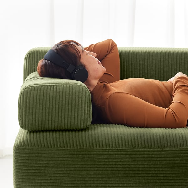 Person relaxing on green JÄTTEBO sofa, wearing headphones, dressed in orange clothes, enjoying leisure time in a cosy setting.