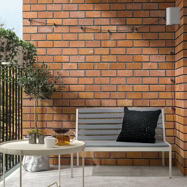 Modern balcony setup with JOLPEN grey outdoor sofa against brick wall, small round side table with tea set, black pillow, and potted plant.