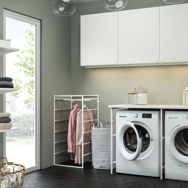 Modern laundry room with white JONAXEL hanging rack holding pink garments, next to white washing machines and cabinets, with a round laundry basket.