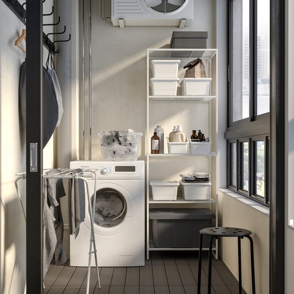 Small, organised laundry area with white JOSTEIN shelves holding containers and bottles. Includes washing machine, drying rack, and stool.