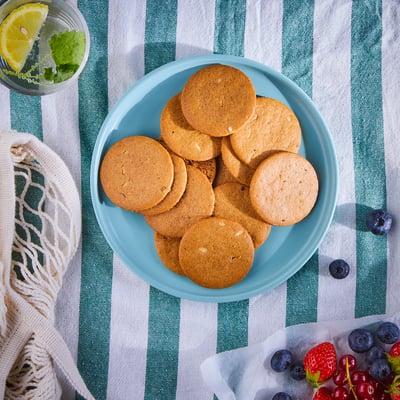 A blue plate of brown ginger cookies sits on a green striped cloth, beside a drink with lemon and mint, and a basket of mixed berries.