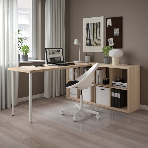 Modern light wood l-shaped desk setup with white chair, laptop, storage, plants, and artwork. Clean, organised, and well-lit.