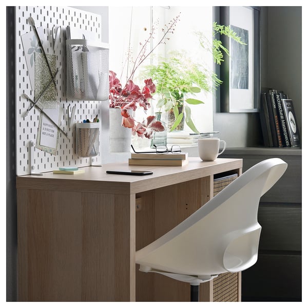Neat home office setup with wooden desk, white swivel chair, organised storage on a wall grid, white mug, books, and a potted plant by the window.