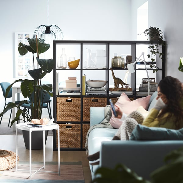 Modern living room with blue KALLAX shelf, plants, and a person on a light blue sofa.