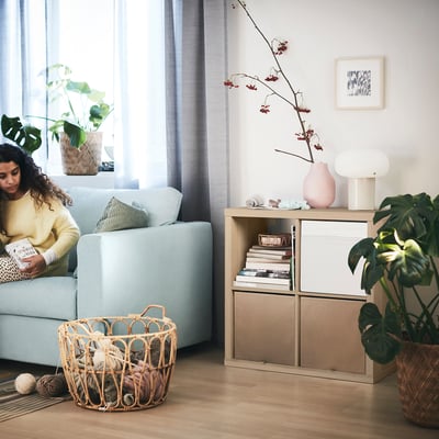 Person reading on blue sofa, KALLAX shelf with books and flowers, plants, yarn basket, blue curtains, wall painting.