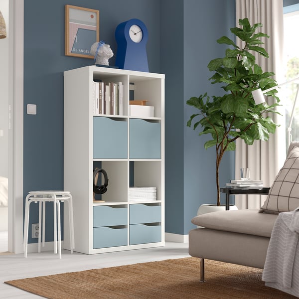 Modern living room with KALLAX bookshelf, blue accents, books, clock. Beige couch, green plant, small stool. Dark blue wall, framed poster.
