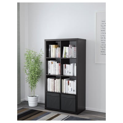 Black KALLAX shelving unit standing, filled with books and decorative items, with a potted plant beside it.
