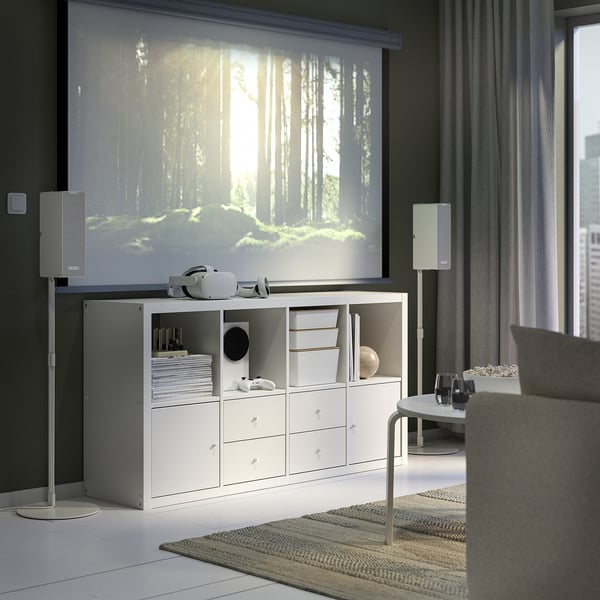 KALLAX shelving with drawers, against a large window, holding books, headphones, and speakers. Partial couch view.